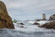 Sea kayaker Leslie Mix in rock garden in moderate swell, near Cape Flattery on the Olympic Coast, with Tatoosh Island lightouse and the Juan de Fuca Pillar behind.