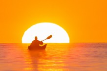 Woman sea kayaker insde the ball off the setting sun, paddles near the Deception Pass Bridge.